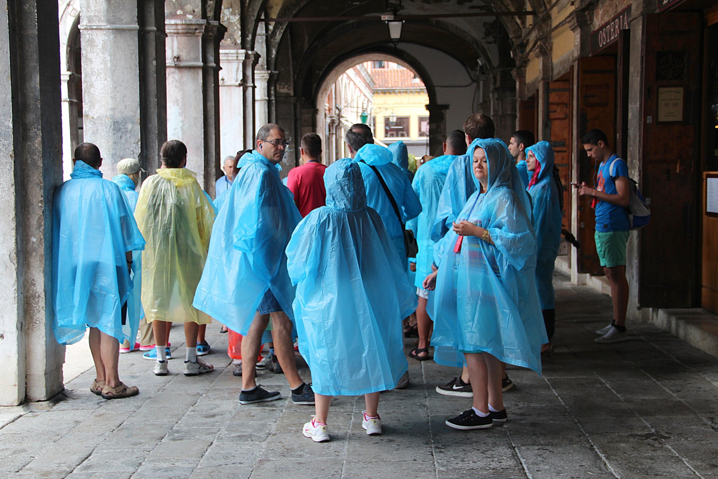 A tour group in matching blue and yellow rain ponchos standing under stone arcades in Venice