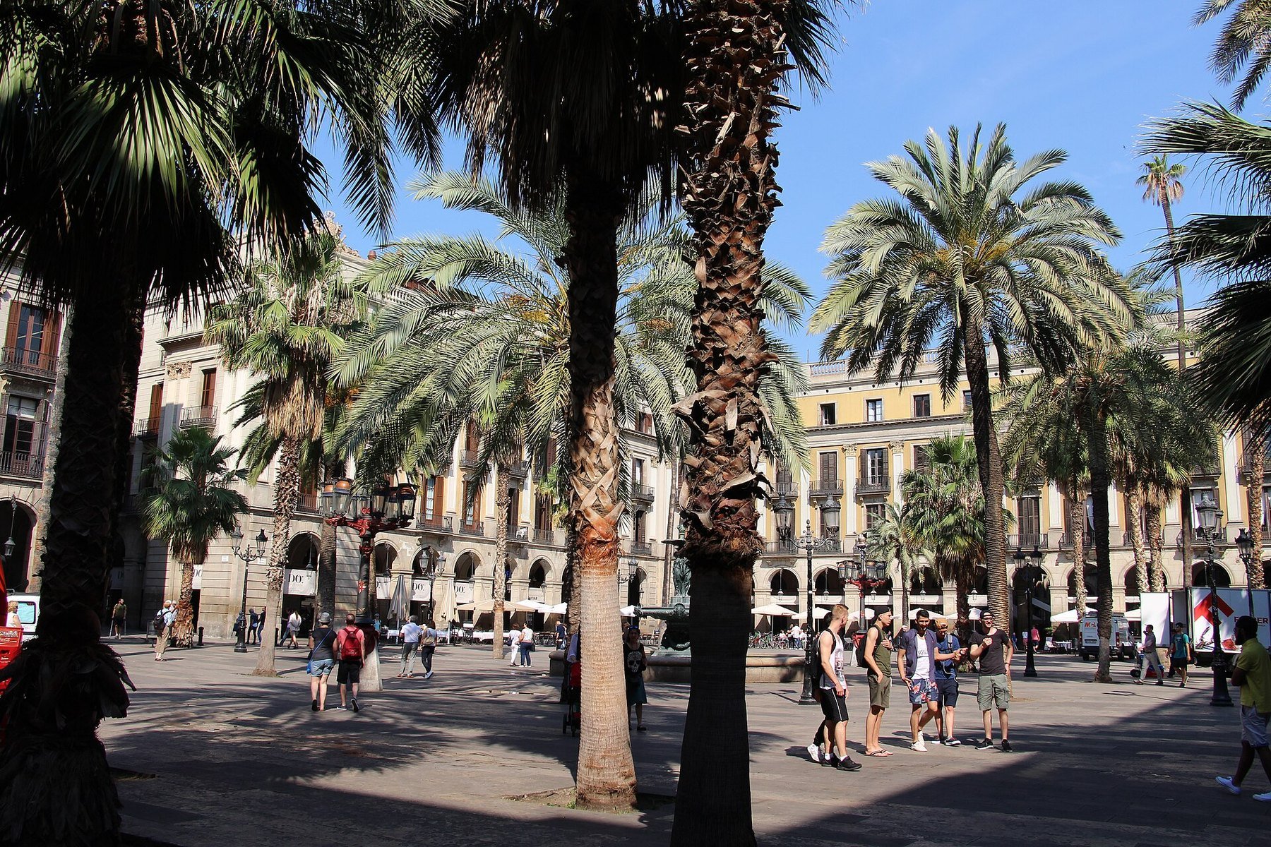 Barselona'da Plaça Reial; palmiye ağaçları, kemerli cepheler ve meydandan geçen ziyaretçiler