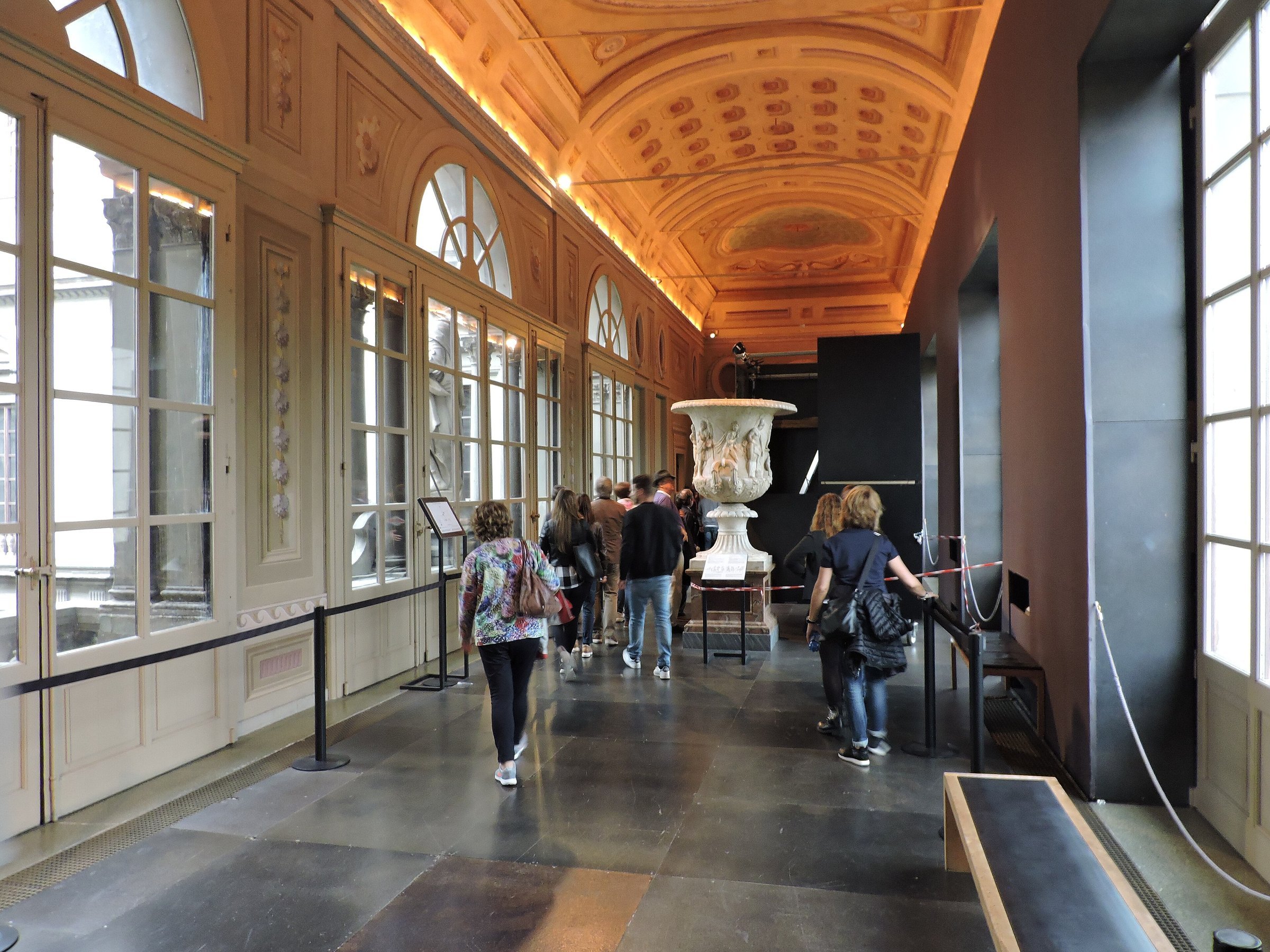 Visitors walking along a long gallery corridor of the Uffizi with a painted ceiling, statues and tall windows