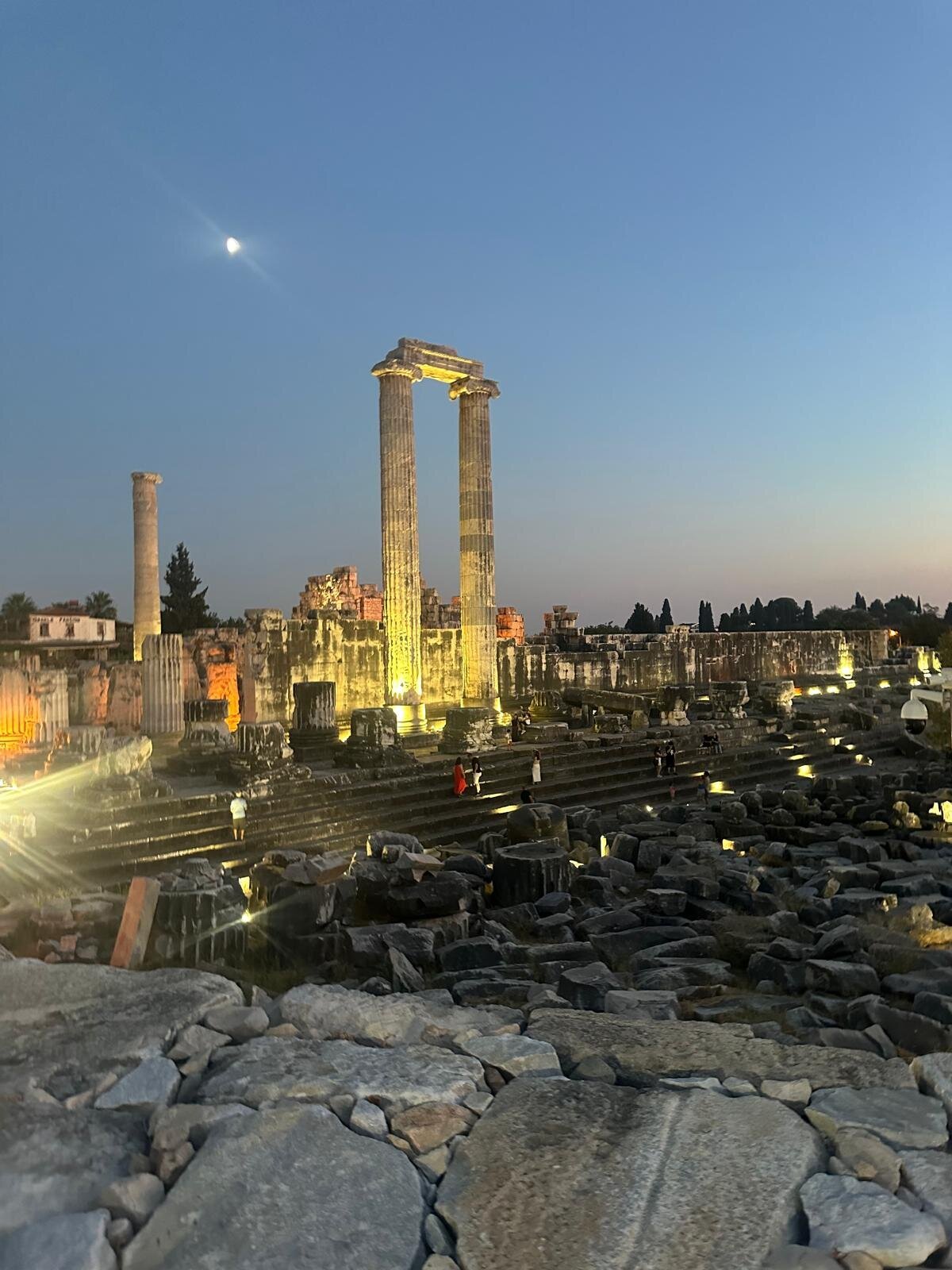 The Temple of Apollo at Didyma floodlit at dusk, with the moon above the two standing columns and visitors on the temple steps.