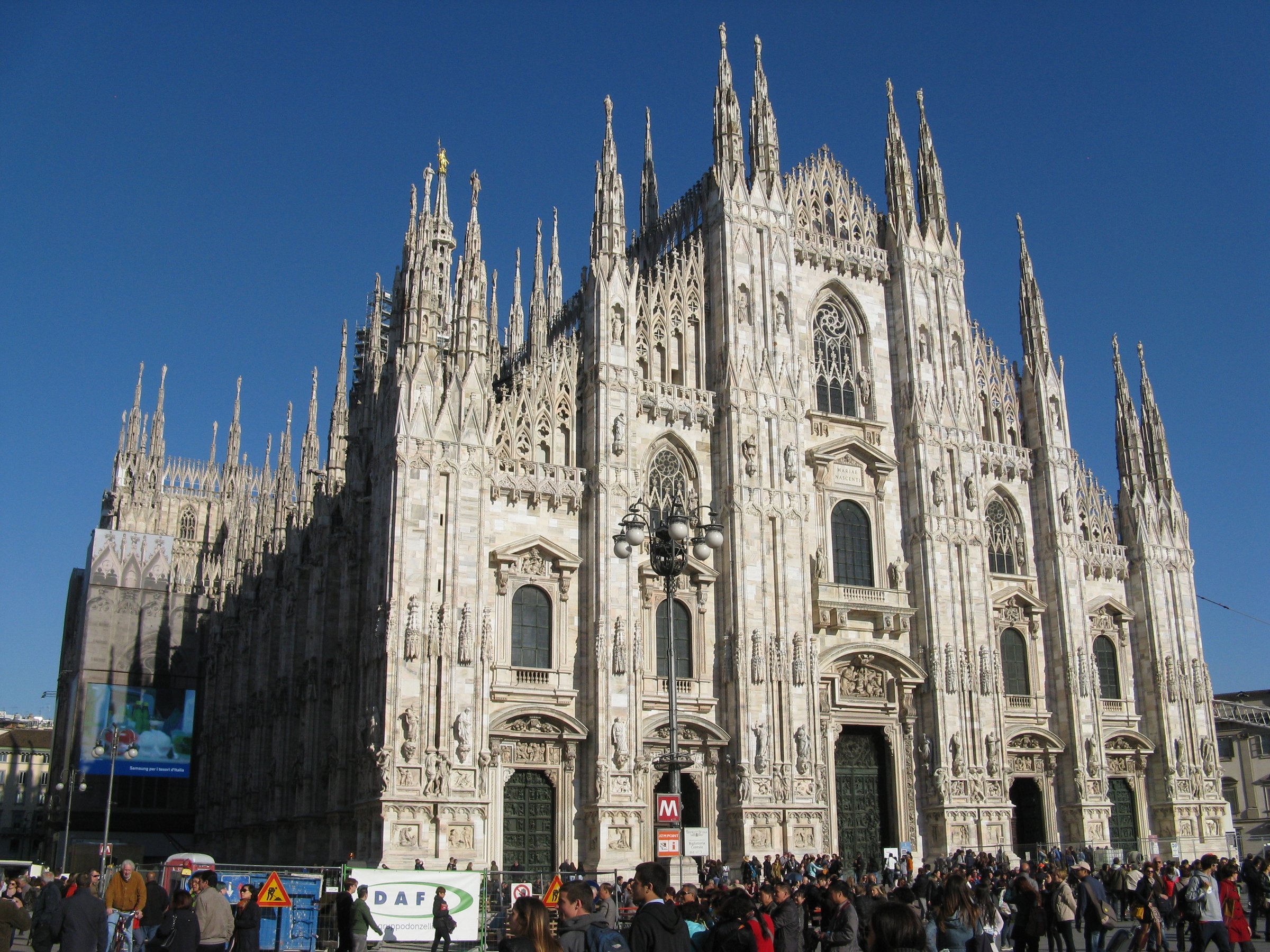 The white marble facade of Milan Cathedral with its spires, and visitors on the square in front