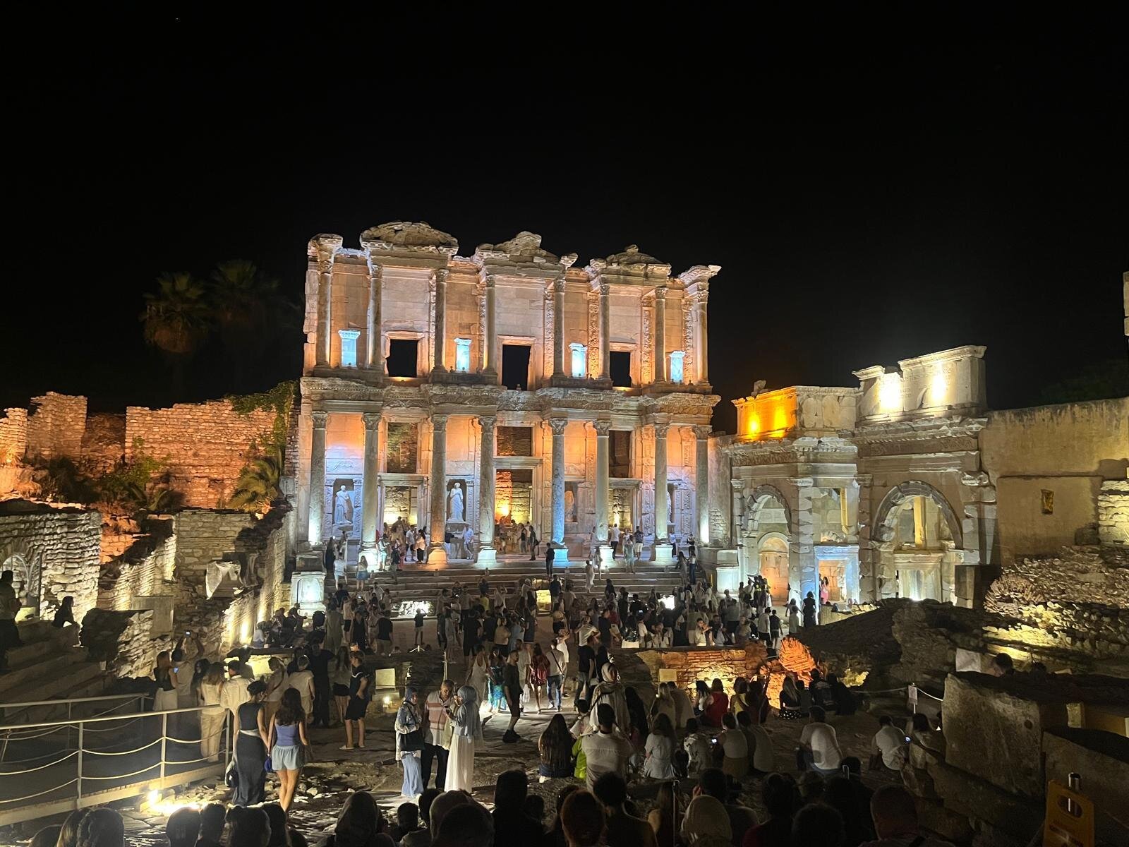 The Library of Celsus at Ephesus lit up at night, with a crowd of visitors gathered on the steps and the terrace in front of it.
