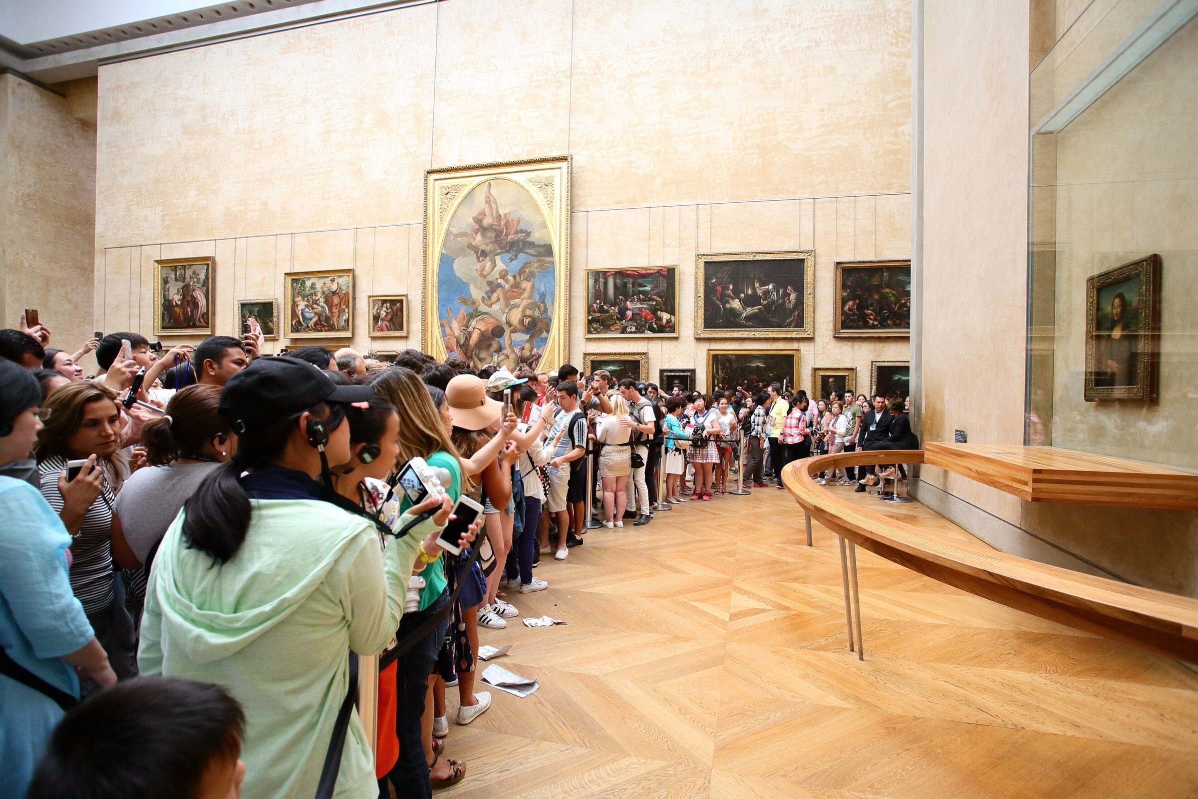 Visitors crowded in front of the Mona Lisa in the Louvre's Salle des États, several holding up phones and one wearing a headset