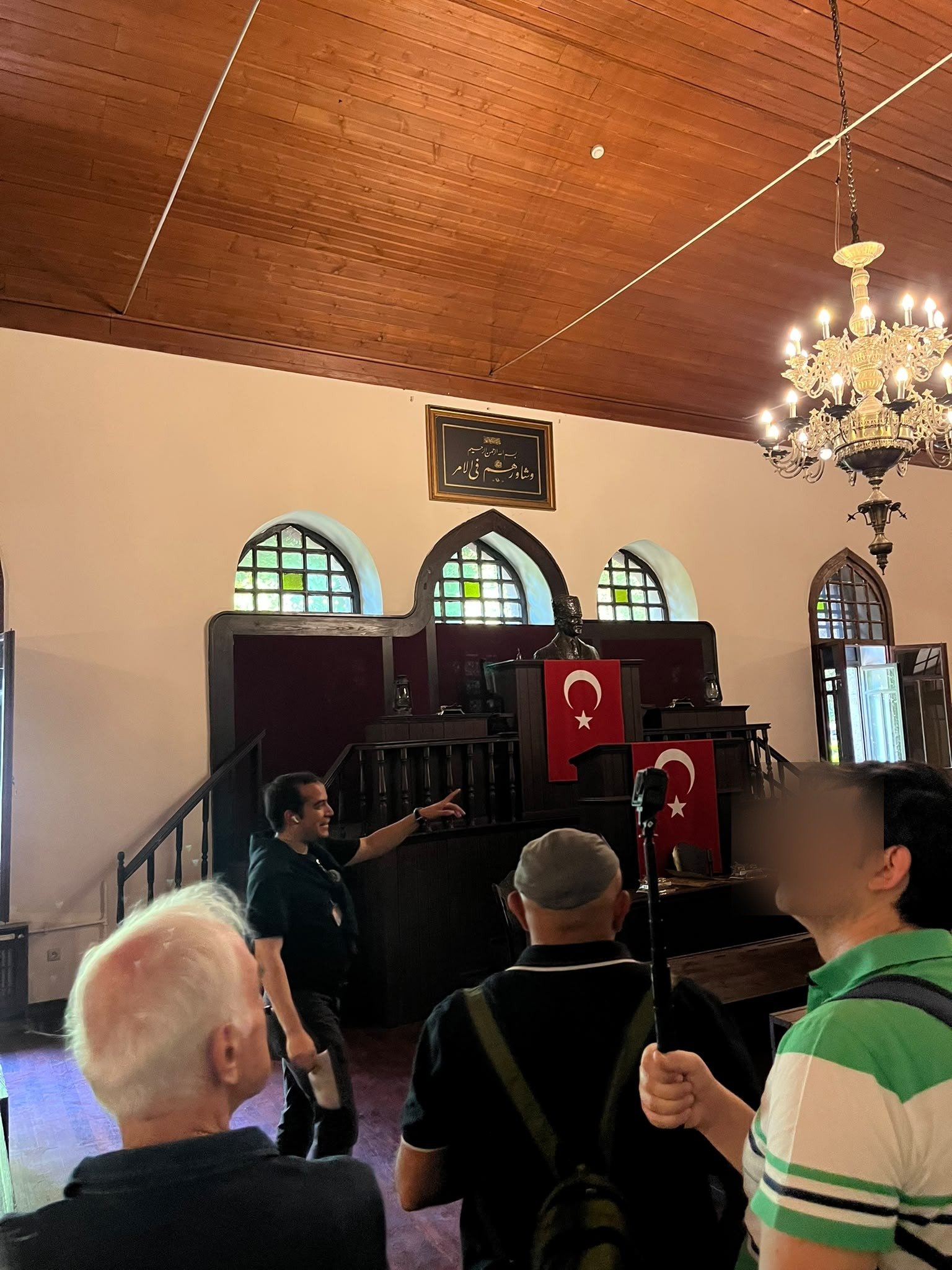Inside the chamber of the First Grand National Assembly building, with a wooden ceiling, arched stained-glass windows and a rostrum flanked by Turkish flags, while the guide points and the group listens.