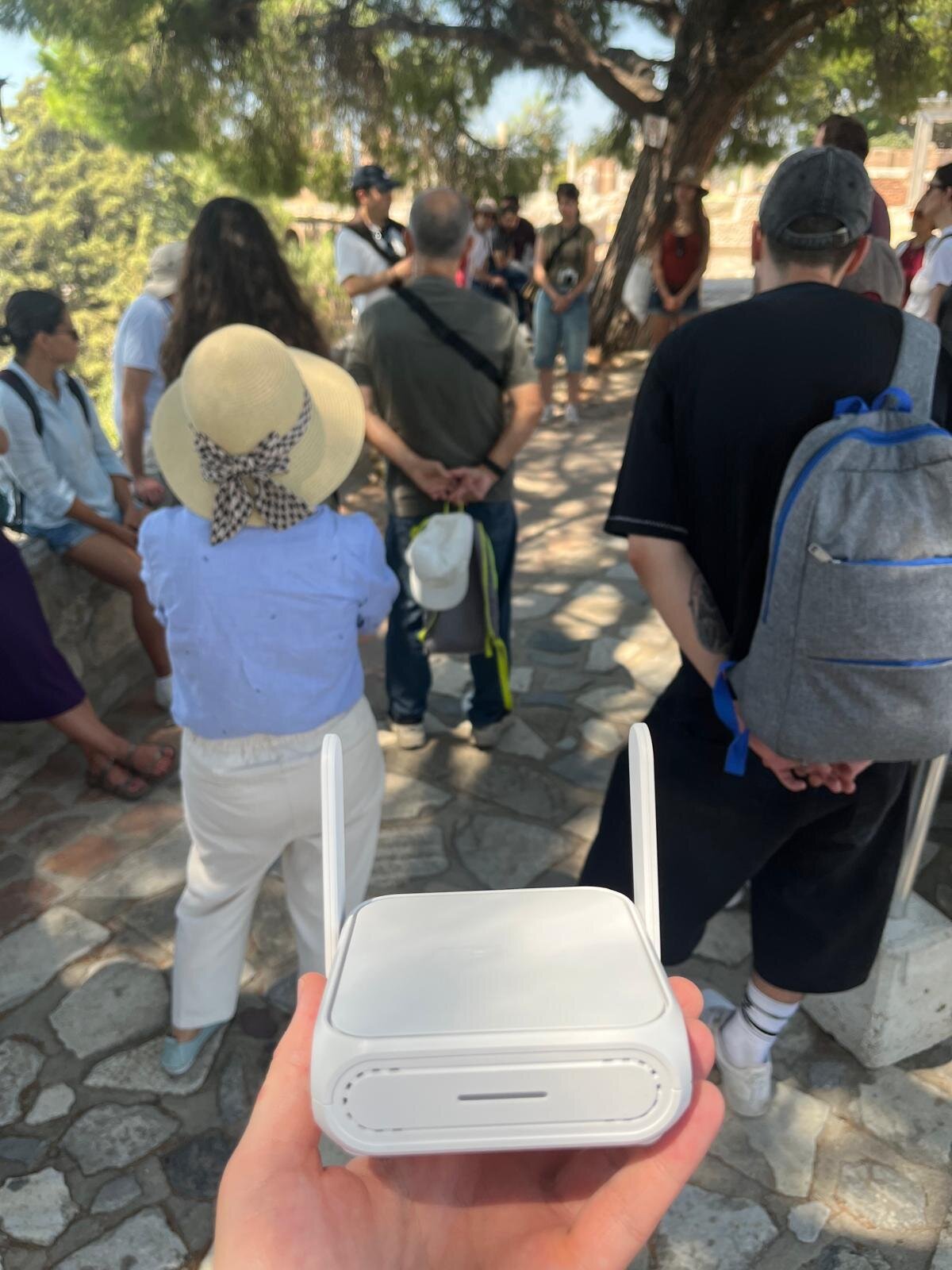 A hand holding a small white portable WiFi router with two antennas, with the tour group standing on a stone path behind it.