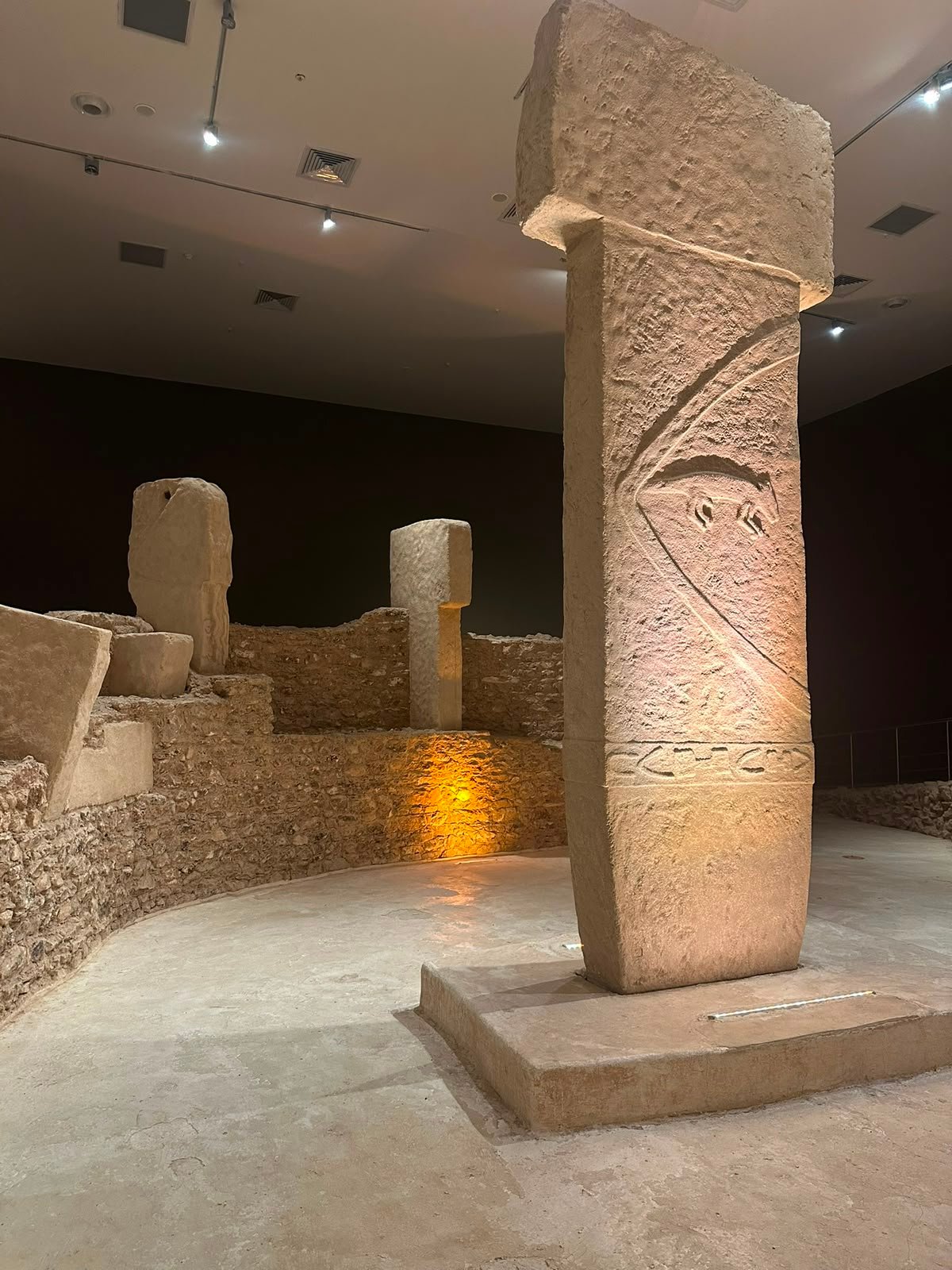 A T-shaped pillar carved with arms and a belt, lit on a plinth in an indoor exhibition hall.