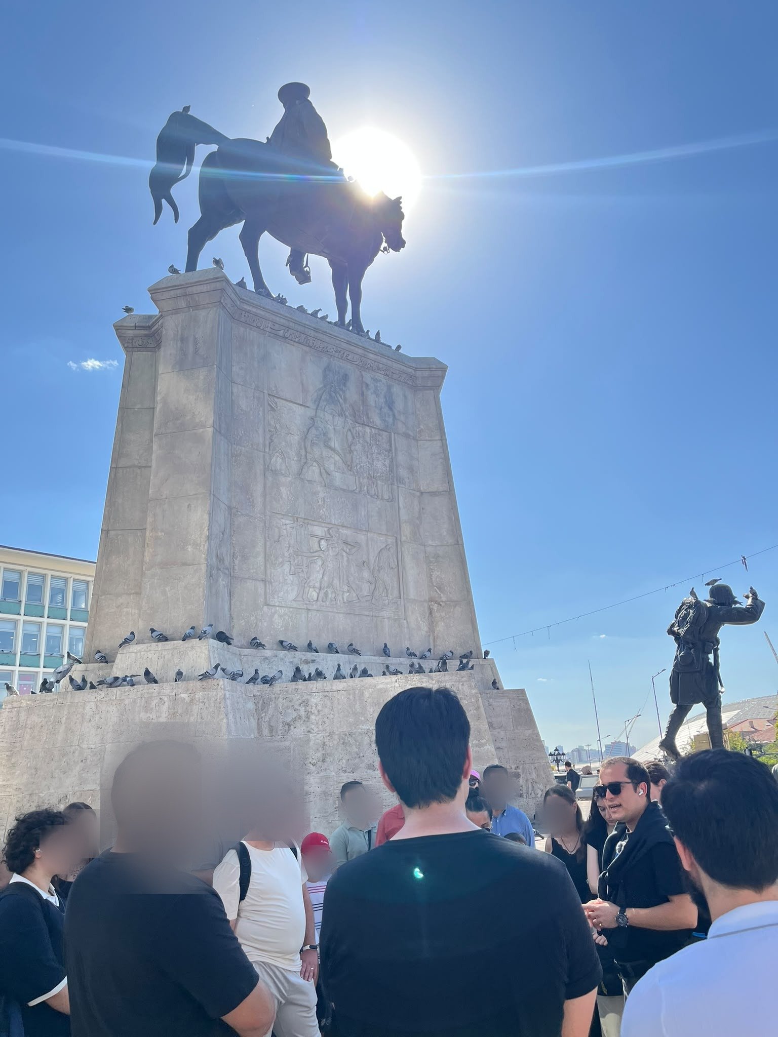 A tour group standing around the guide in Ulus Square, below the equestrian Victory Monument, with the sun behind the statue.