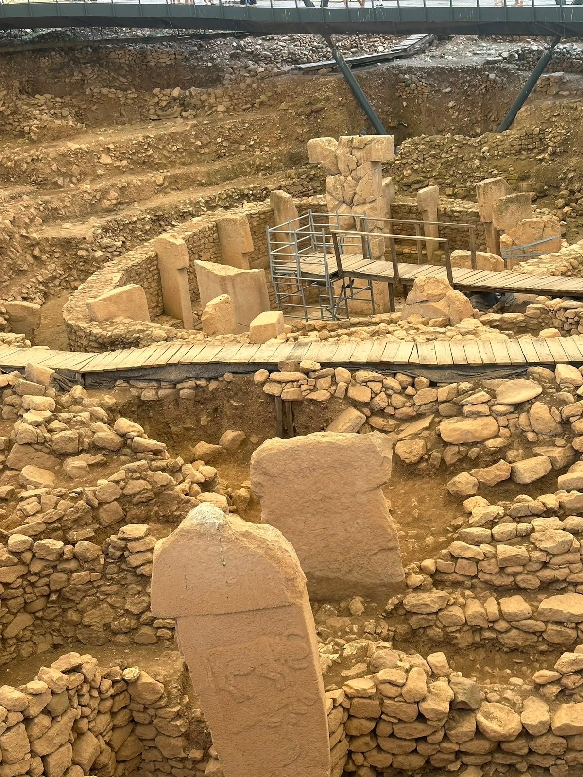 Several excavated enclosures with T-shaped pillars, crossed by wooden walkways, with visitors on the elevated bridge above.