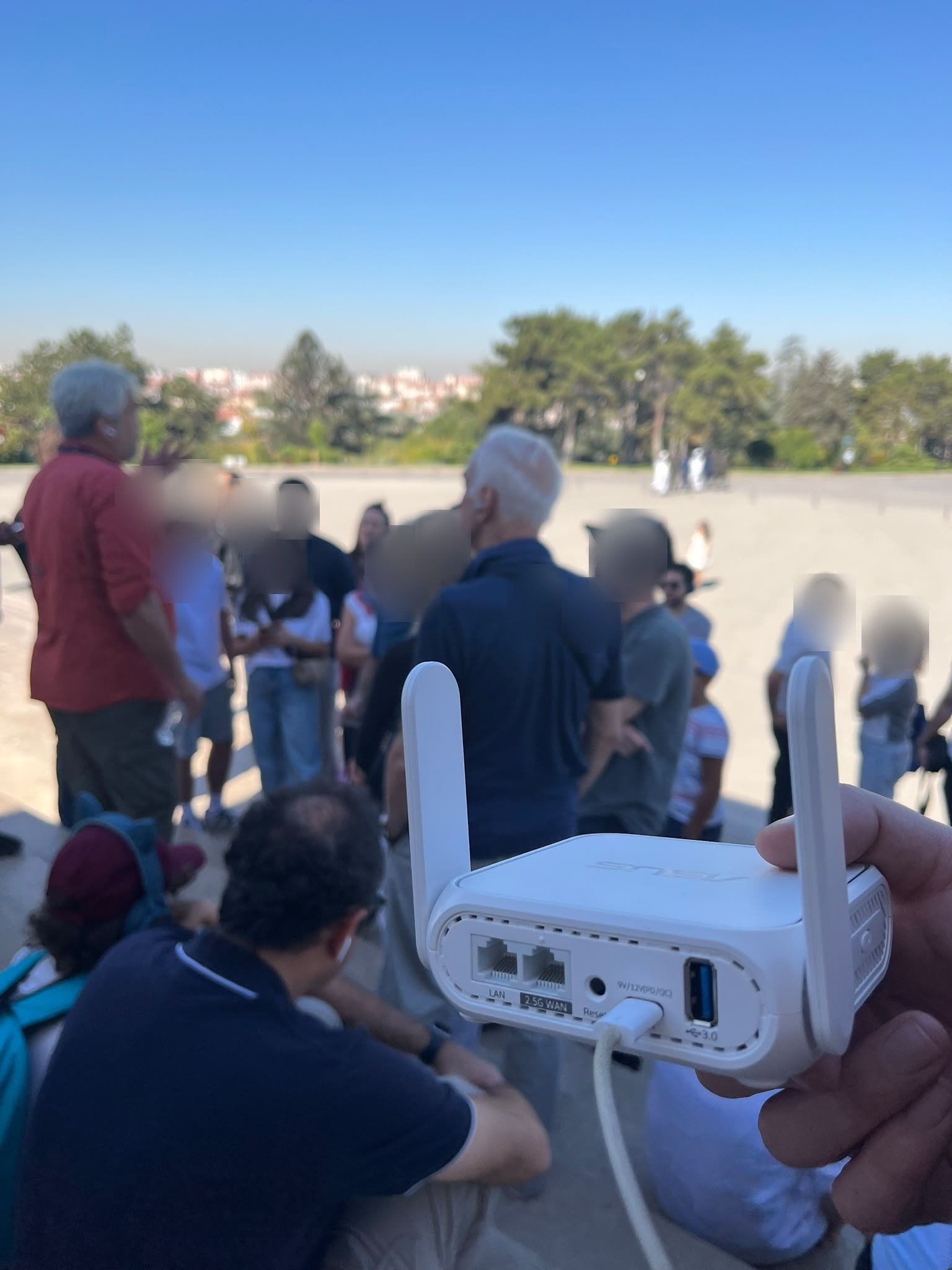 A hand holding a small white portable WiFi router with two antennas, with the tour group gathered around the guide on open ground behind it.
