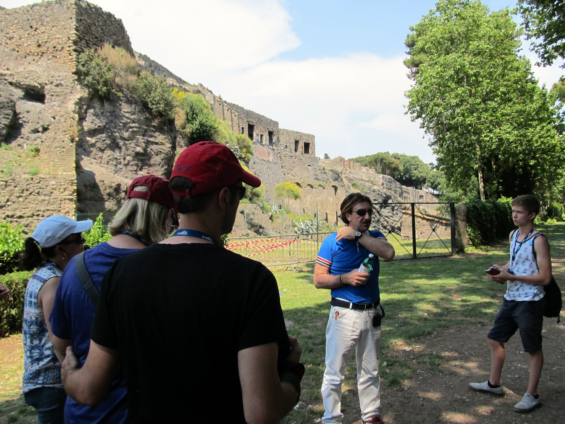 A guide talking to a small group of visitors on a grassy path among the ruins of Pompeii