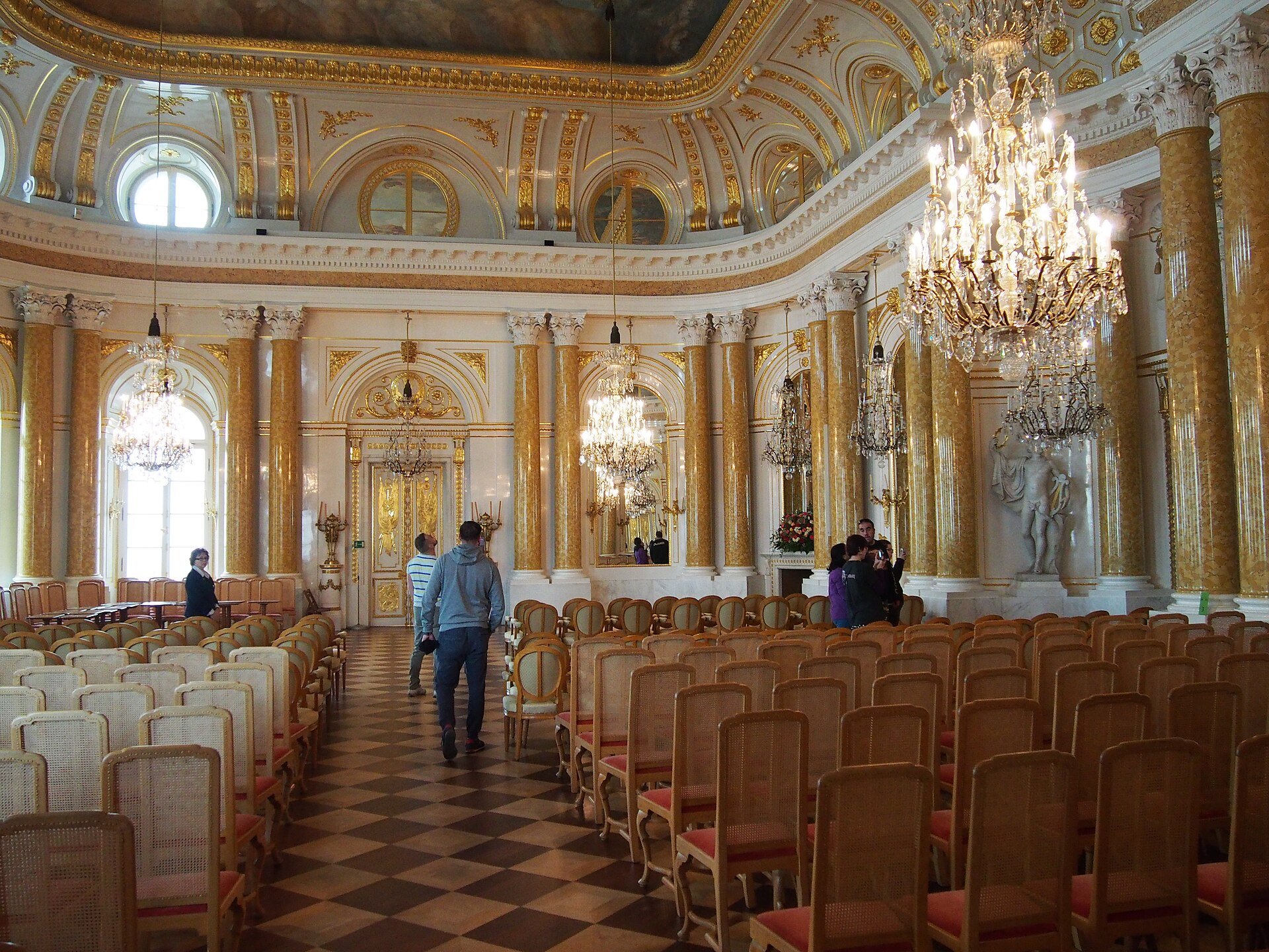 The Great Hall of the Royal Castle in Warsaw, with gilded columns, chandeliers and rows of chairs, and a few visitors walking through
