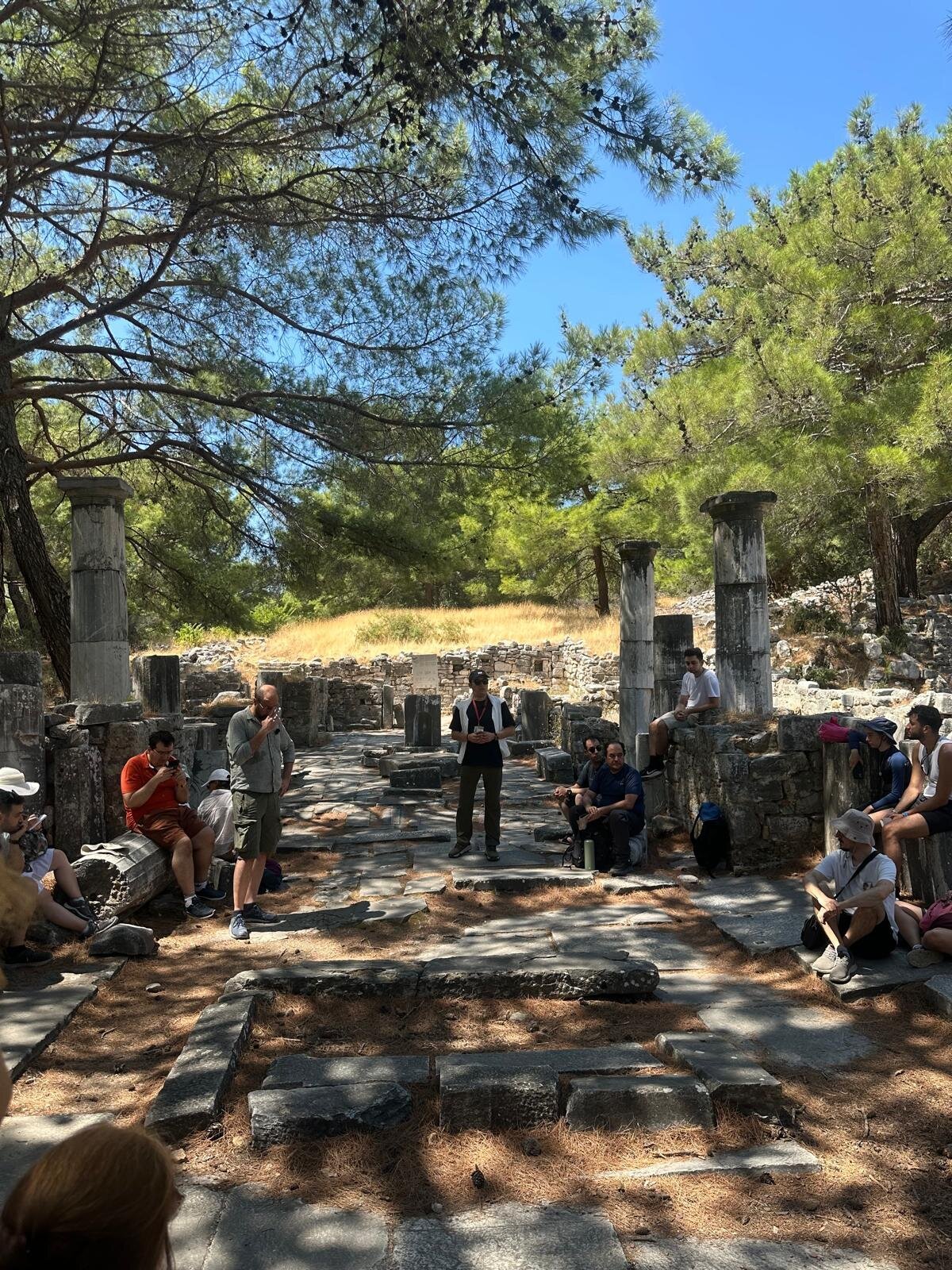 A tour group seated on fallen stones inside the ruined walls of an old cathedral, with the guide standing in the middle and pine trees overhead.