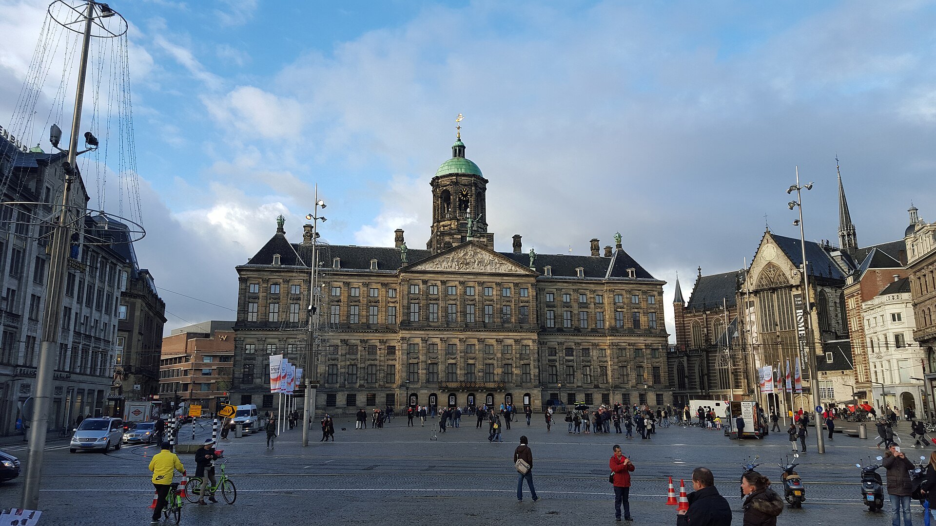 The Royal Palace on Dam Square in Amsterdam, a wide classical facade with a domed tower, with the Nieuwe Kerk to the right and people crossing the square