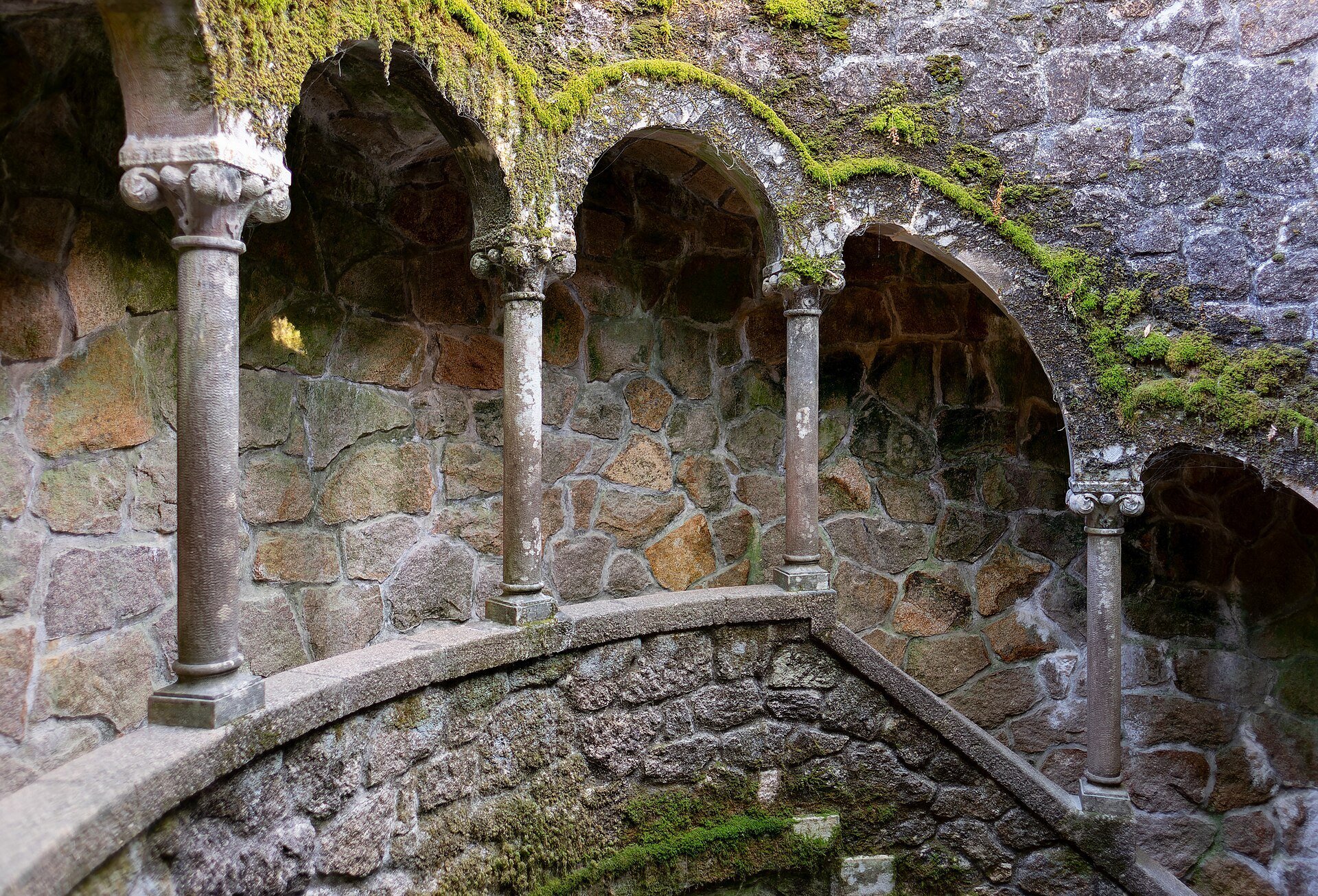 Looking down the spiral staircase of the Initiation Well at Quinta da Regaleira, with moss-covered stone arches and columns