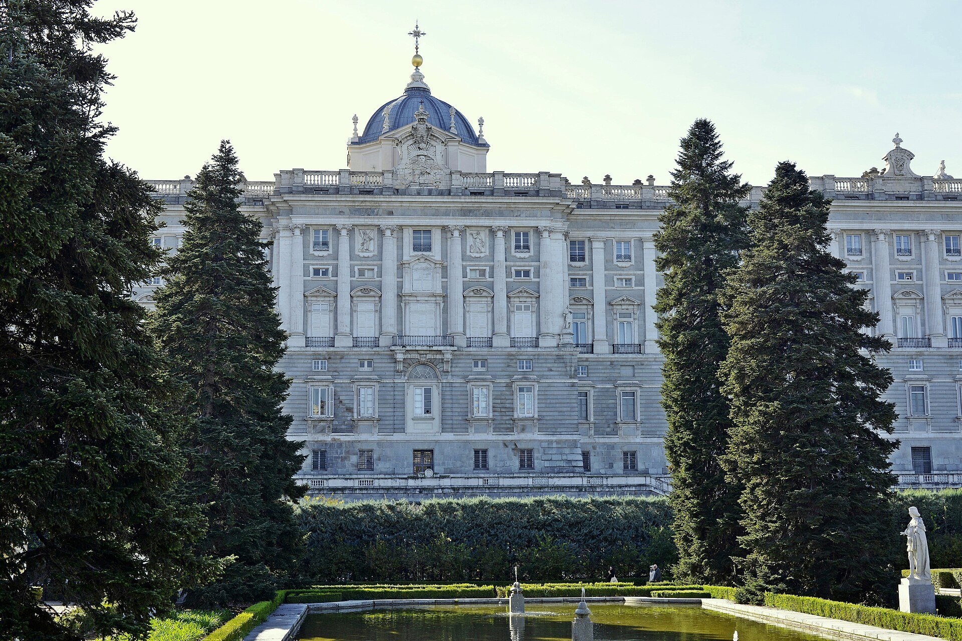 The north facade of the Royal Palace of Madrid seen across the Sabatini Gardens, with tall conifers and a pond in front