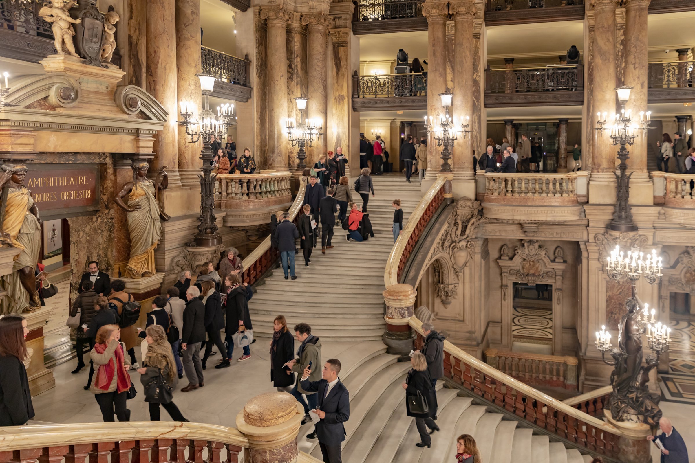Visitors on the marble Grand Staircase of the Palais Garnier, lit by bronze candelabra