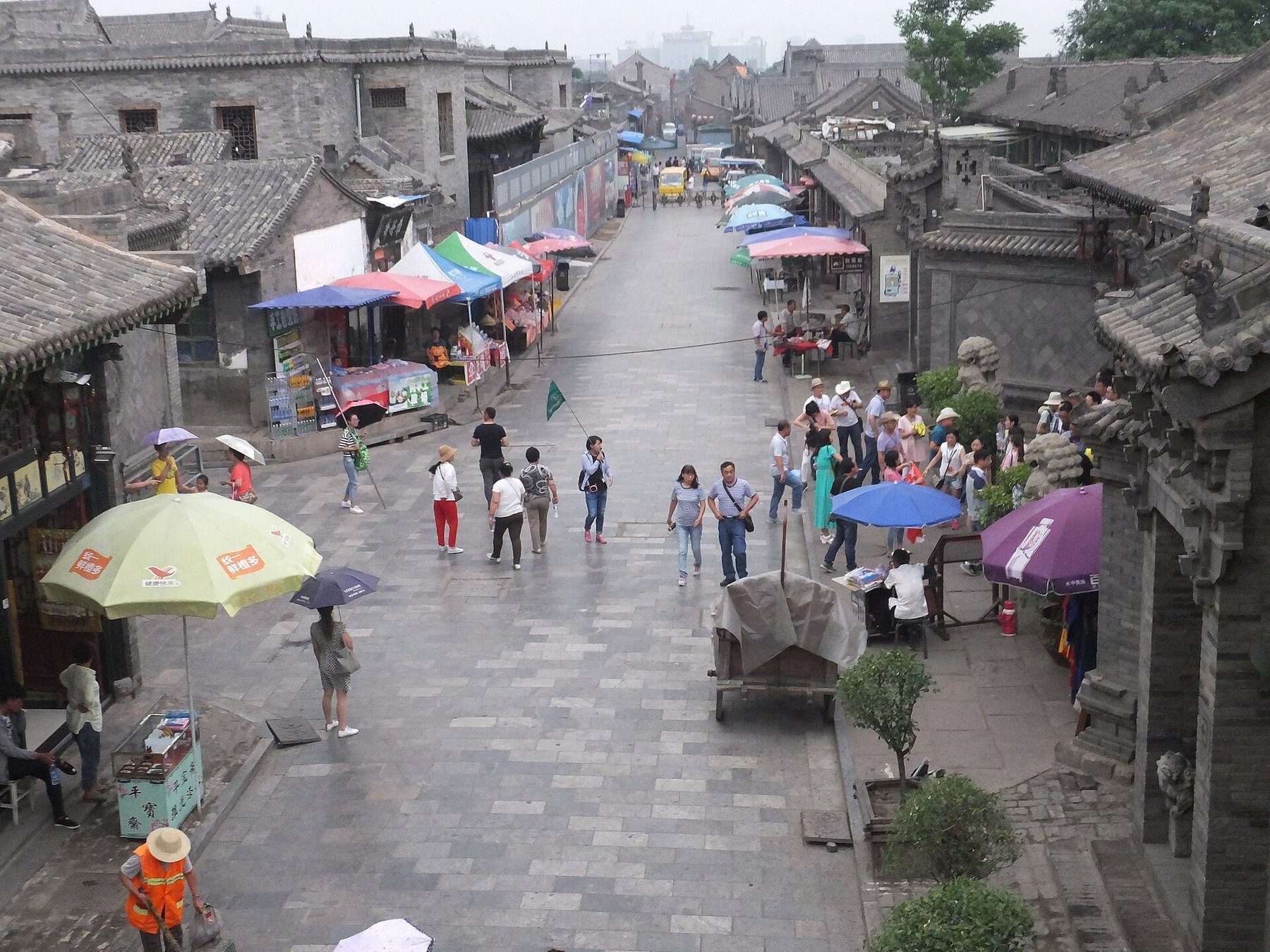 A paved street in Pingyao Ancient City lined with grey brick shops, umbrellas and walking visitors